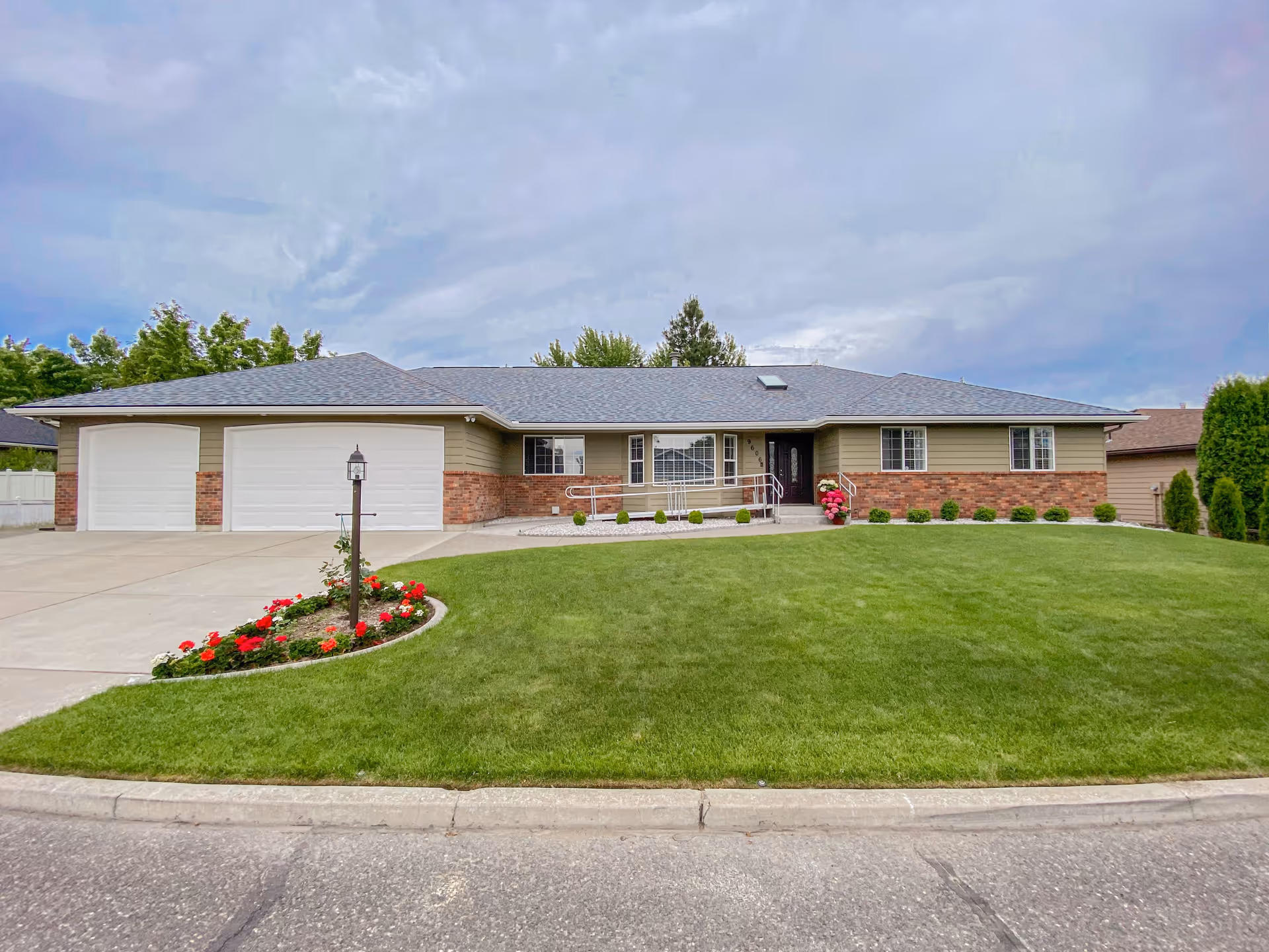 Single-story residential building with a three-car garage, beige siding with brick accents, a well-maintained green lawn, a flower bed with red and white flowers, and a paved driveway. The entrance features a ramp with handrails and a black front door under a cloudy sky.