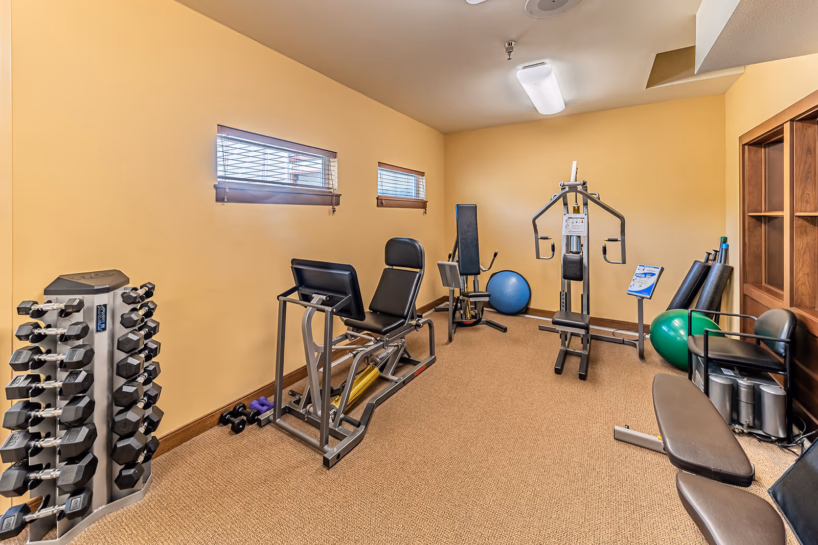 A small fitness room with beige walls and carpeted floor featuring various exercise equipment including a leg press machine, a multi-function weight machine, a rack of dumbbells, exercise balls, foam rollers, and a bench. Two small windows with blinds allow natural light into the room.