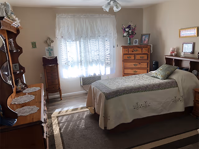 A cozy bedroom with a single bed covered in a floral quilt and a pillow. The room has wooden furniture including a dresser with a mirror, a chest of drawers, a small cabinet, and a nightstand. There are lace curtains on the window letting in natural light, and decorative items such as framed pictures, a vase with flowers, and a teddy bear on the furniture.
