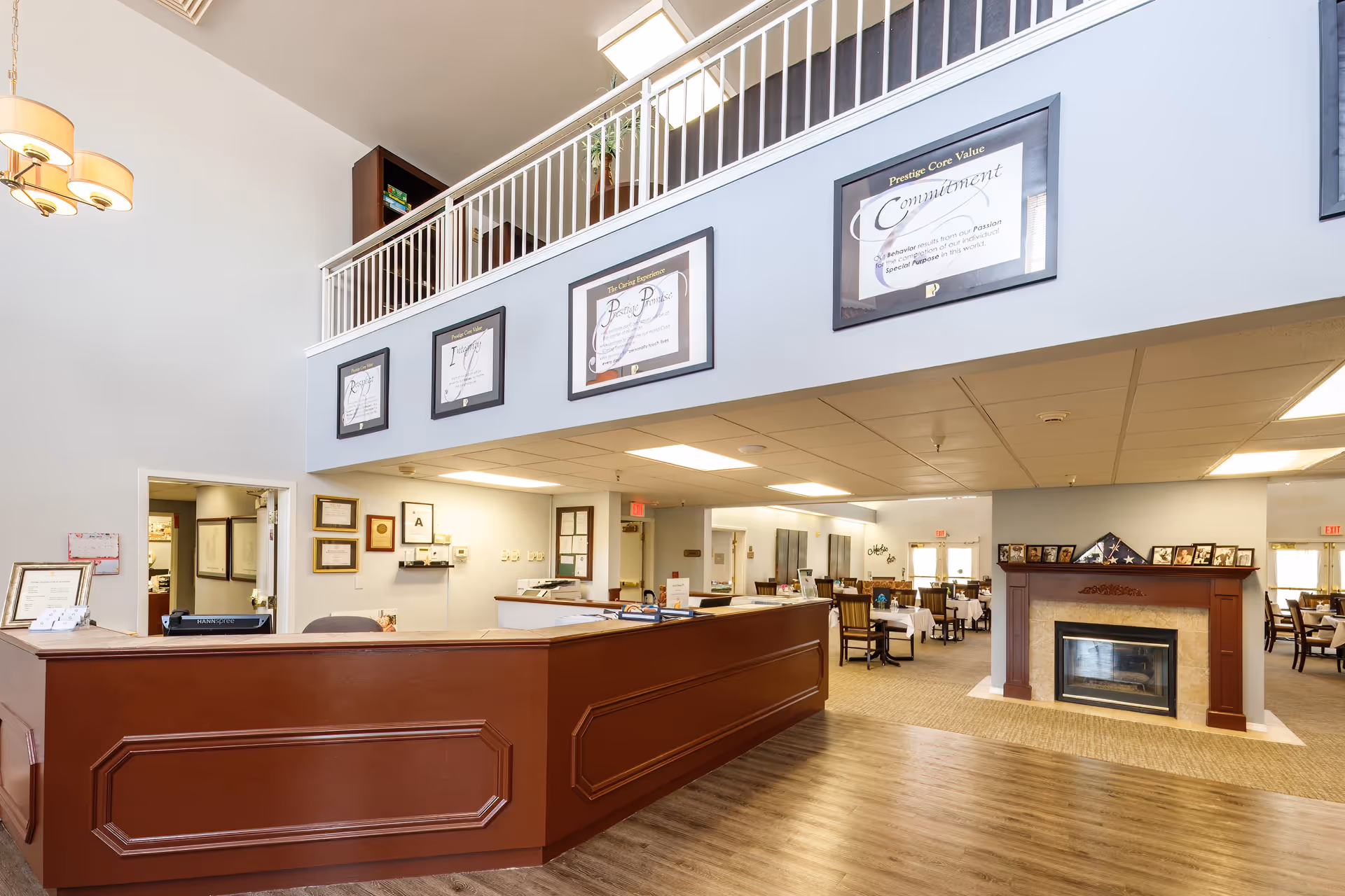 Interior view of a senior living facility reception area with a large wooden front desk, framed certificates and awards on the walls, a fireplace with framed photos on the mantel, and a dining area with tables and chairs in the background.