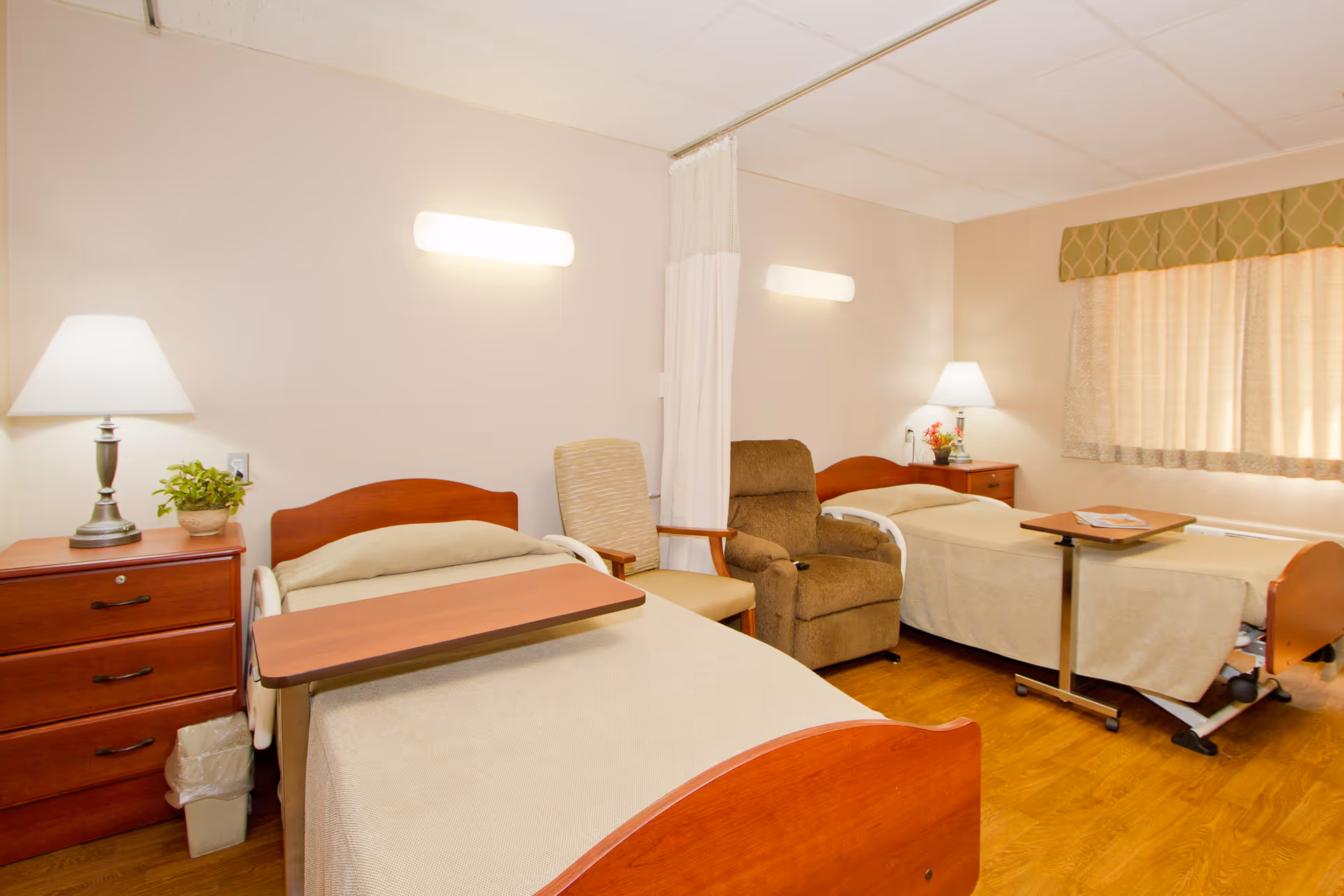 A well-lit senior living bedroom with two hospital-style beds, each with a wooden headboard and beige bedding. Between the beds are two chairs, one beige and one brown recliner. Each bed has an overbed table. There are two wooden nightstands with lamps and a small plant on one. A window with beige curtains and a green valance is on the right wall. The room has light-colored walls and a wood floor.
