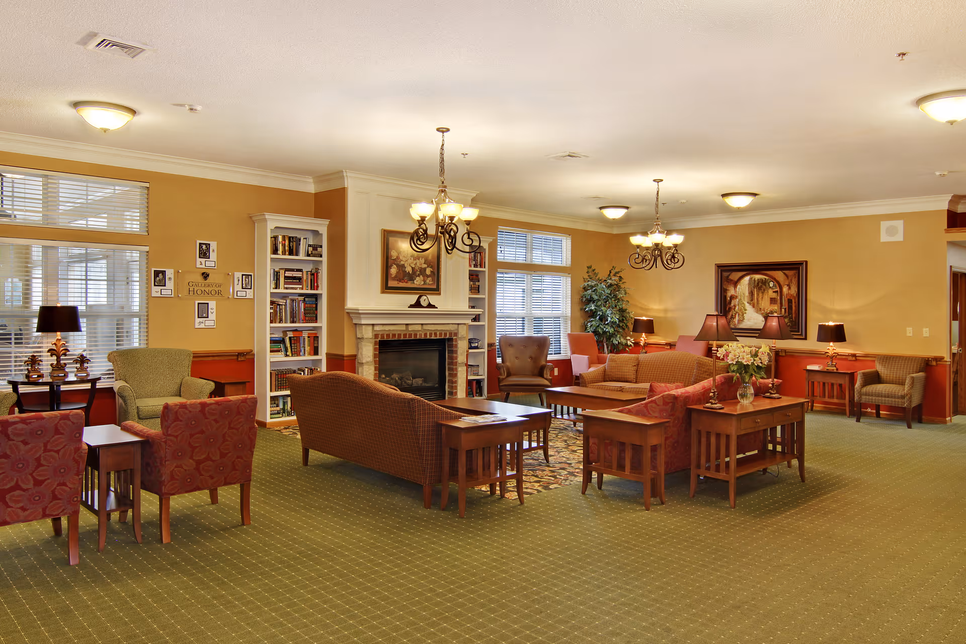 A spacious and warmly lit living room area in a senior living facility with multiple seating arrangements including armchairs and sofas around wooden coffee tables. There is a fireplace with a painting above it, bookshelves, table lamps, and large windows with blinds allowing natural light to enter. The walls are painted in warm tones and the carpet is green with a subtle pattern.