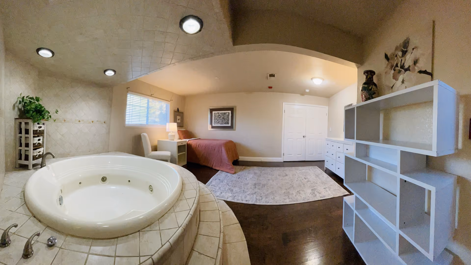 Spacious bedroom with a built-in round whirlpool tub in the foreground and a bed, dresser, and shelving in the background.