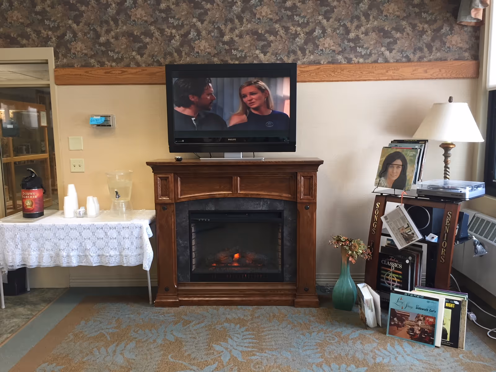 A cozy common room with a wall-mounted TV above a wooden electric fireplace, a table with coffee cups on the left and a lamp and shelf of records on the right.