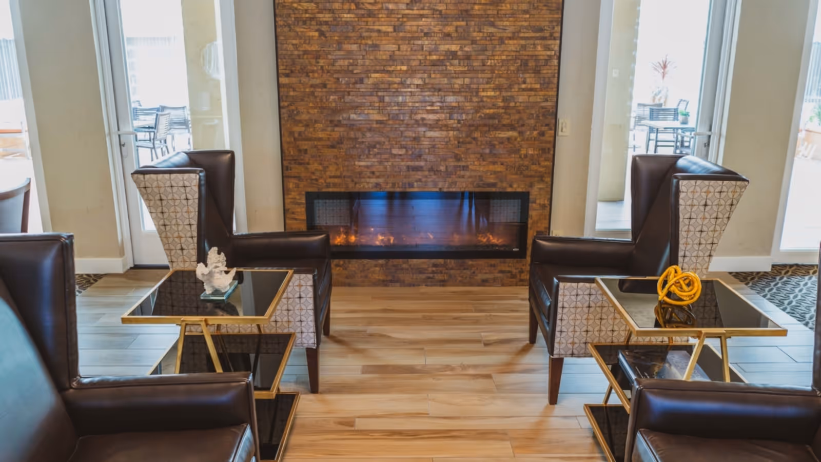 A cozy seating area with four leather armchairs arranged around two modern glass and gold side tables, positioned in front of a wall-mounted electric fireplace with a brick surround. Large glass doors on either side lead to an outdoor patio with additional seating.