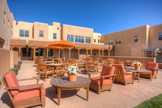Outdoor patio area at The Montecito of Santa Fe featuring multiple wooden tables and chairs with orange cushions, some tables shaded by orange umbrellas, and potted plants on tables. The patio is surrounded by beige stucco buildings under a clear blue sky.