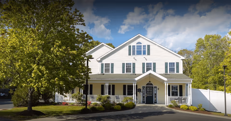 A large two-story white building with green shutters and a covered front porch, surrounded by trees and landscaping under a partly cloudy sky.