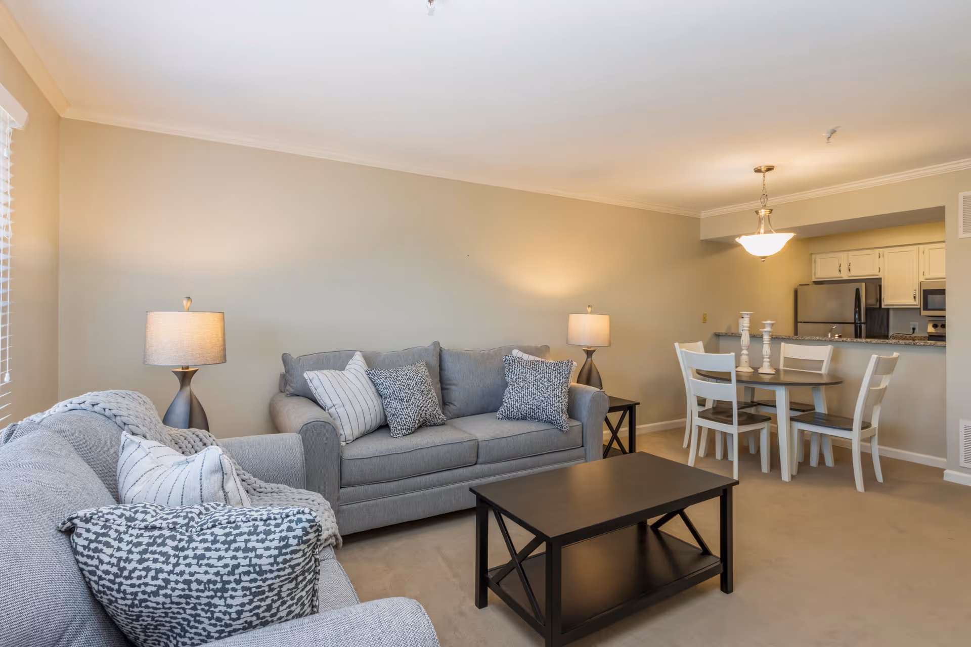 A cozy living room with two gray sofas adorned with patterned pillows and a gray throw blanket. There are two matching table lamps on either side of the sofas. A dark wooden coffee table is centered in front of the sofas. In the background, there is a dining area with a round table and four white chairs, and a kitchen with white cabinets and stainless steel appliances.