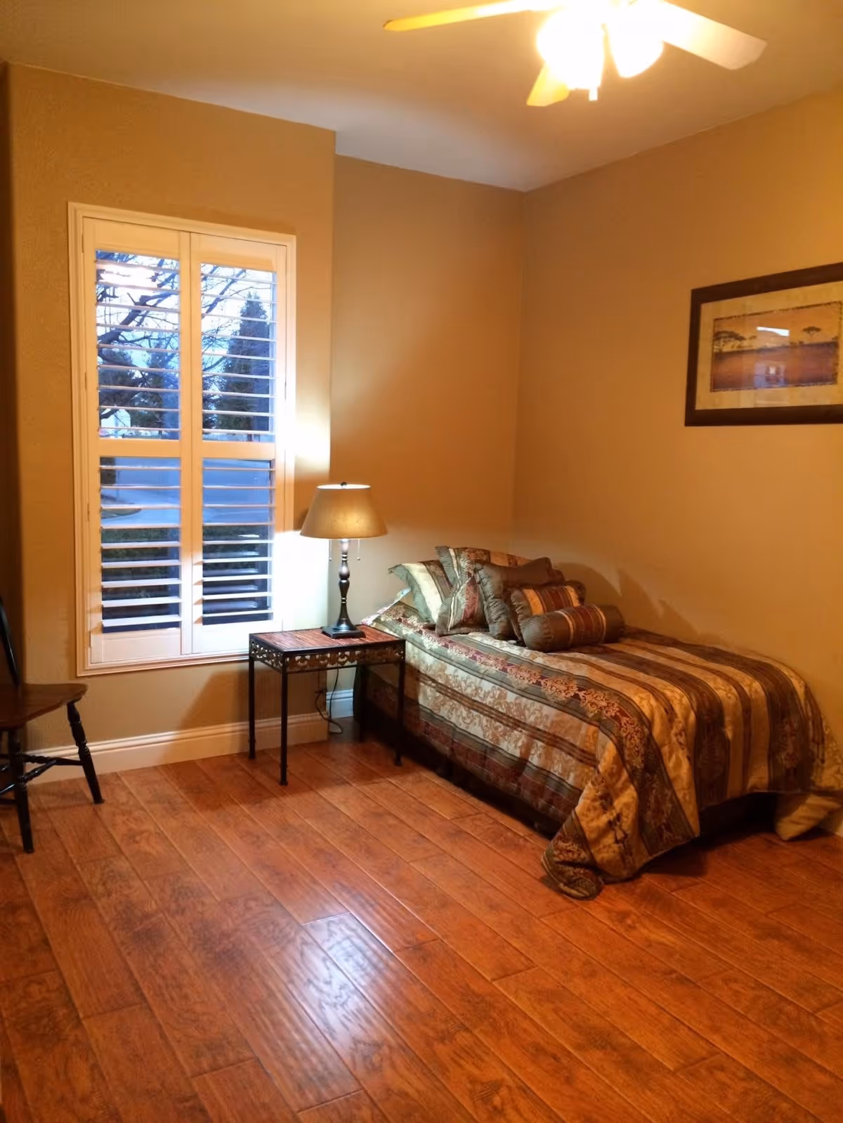 A cozy bedroom with a single bed covered in a patterned bedspread and multiple pillows. Next to the bed is a small side table with a lamp. There is a window with white shutters on the left wall and a framed picture hanging on the right wall. The floor is wooden and there is a wooden chair partially visible on the left side.