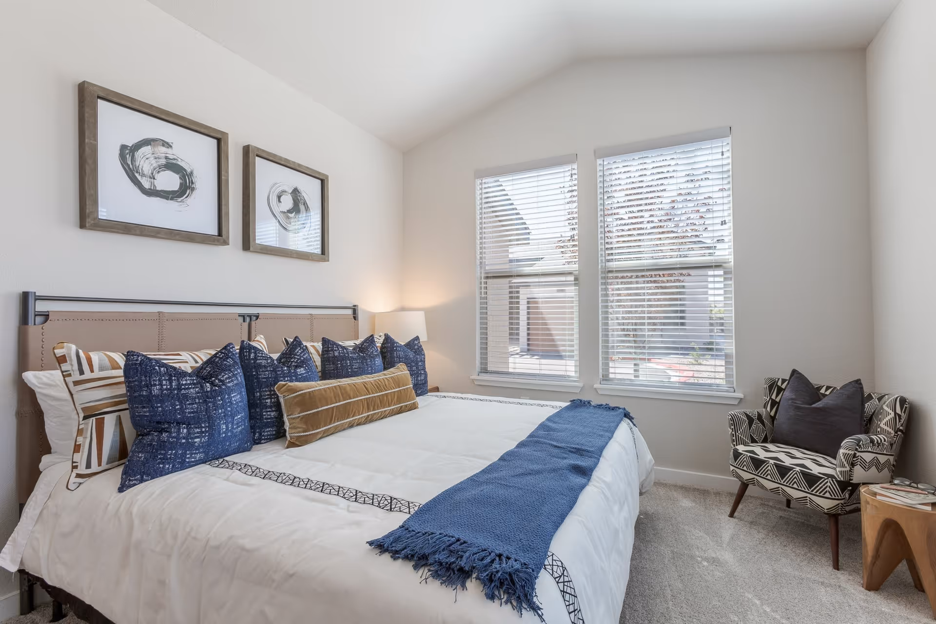 A bright bedroom with a large bed featuring multiple pillows in blue, brown, and white patterns. The bed has a white comforter with a blue throw blanket at the foot. Two abstract framed artworks hang above the bed. There are two windows with white blinds letting in natural light. A patterned armchair with a dark pillow sits in the corner next to a small wooden side table with magazines.
