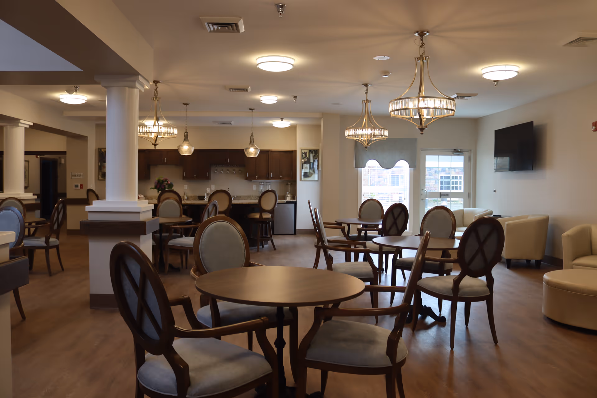 Bright communal dining and lounge area with round tables, upholstered chairs, chandeliers and a kitchenette in the background.