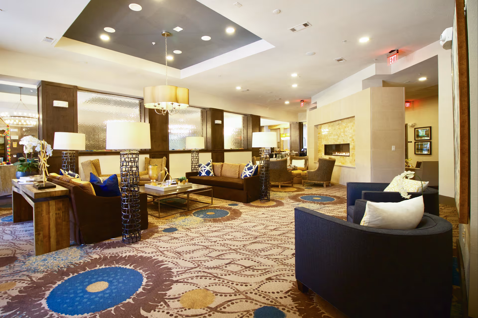 A spacious and well-lit living room area in a senior living facility featuring multiple seating arrangements including brown and blue armchairs and sofas with decorative pillows. The room has patterned carpet with blue and beige circular designs, several modern floor lamps, a hanging ceiling light, and a built-in fireplace on the right wall. The walls have dark wood paneling with frosted glass windows, and there are exit signs visible in the background.