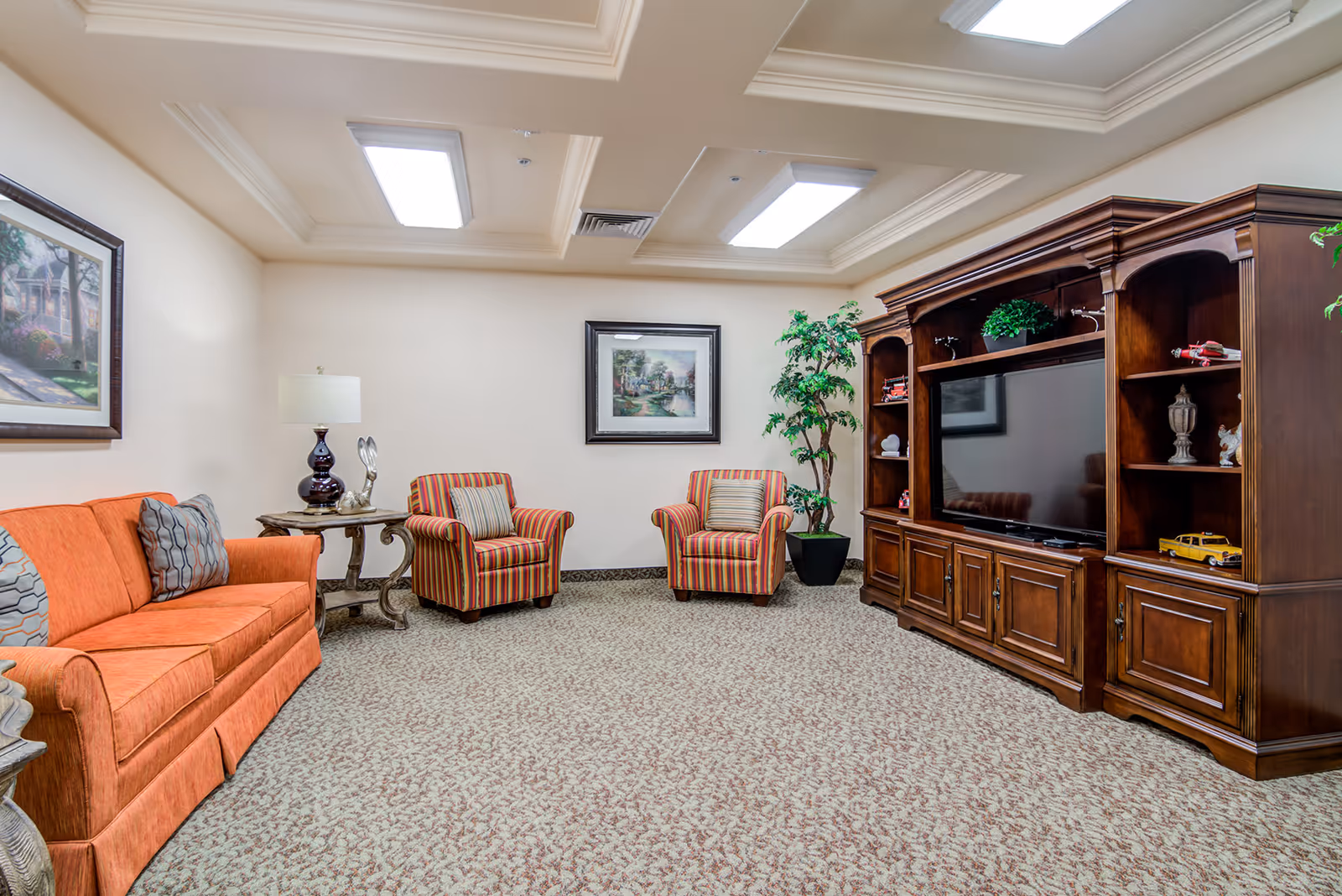 A cozy living room with an orange sofa, two striped armchairs, a wooden entertainment center with a large flat-screen TV, a potted plant, and framed artwork on the walls. The room has a patterned carpet and recessed ceiling lights.