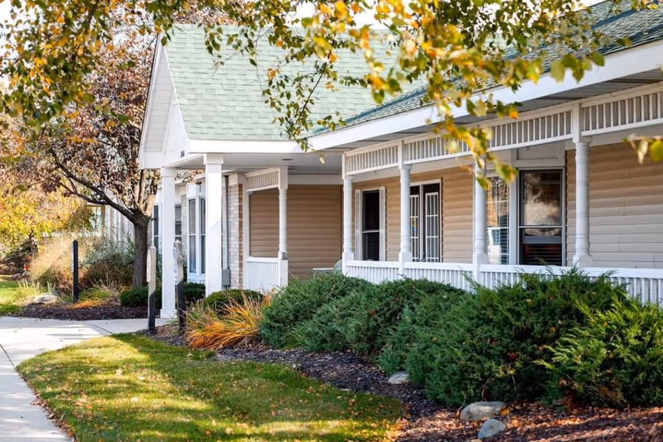 Exterior view of a senior living facility building with beige siding, white columns, and a green roof. There is a covered porch with white railings and several windows. The surrounding area has green bushes, trees with autumn leaves, and a sidewalk leading to the entrance.