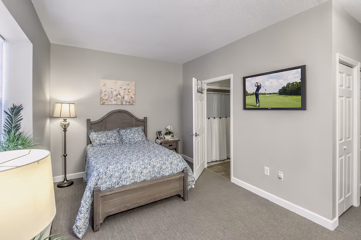A furnished bedroom with a wooden bed, nightstand and lamp, a wall-mounted TV, and an open door to a bathroom.