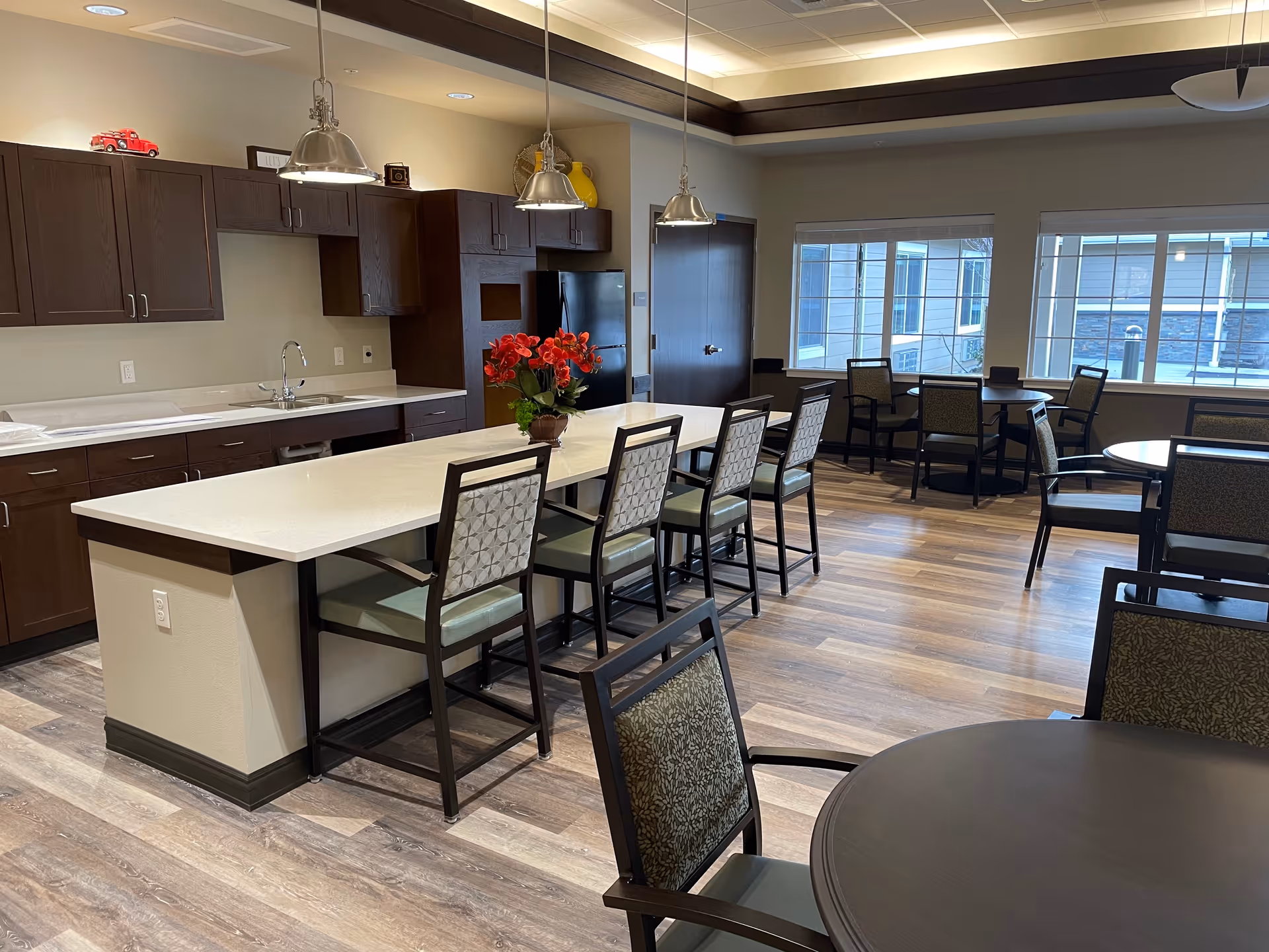 A spacious dining area with a modern kitchen island featuring a white countertop and six chairs. The kitchen has dark wood cabinets, a sink, and a black refrigerator. There are three pendant lights hanging above the island. In the background, there are multiple round tables with chairs near large windows letting in natural light. The floor is wood-patterned, and the ceiling has recessed lighting.