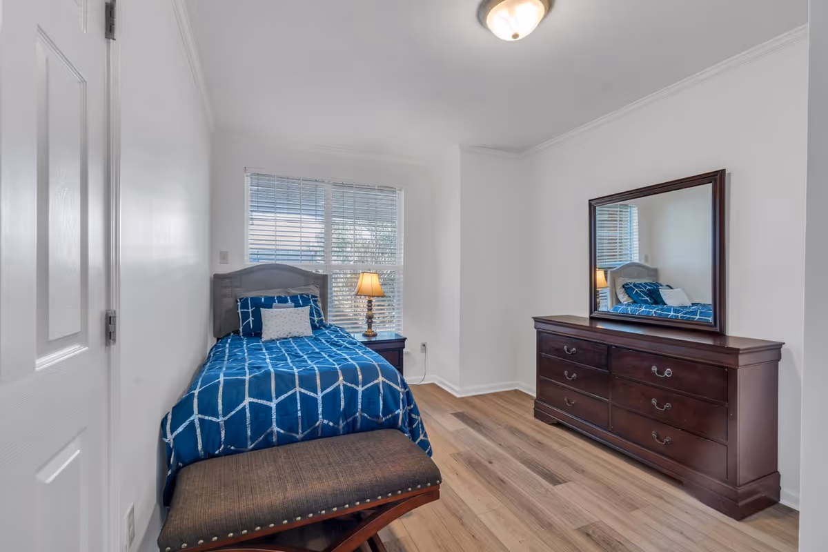 A small bedroom with a single bed covered in a blue and white patterned comforter, a nightstand with a lamp next to the bed, a large wooden dresser with a mirror above it, and a cushioned bench at the foot of the bed. The room has white walls, a window with blinds, and wooden flooring.