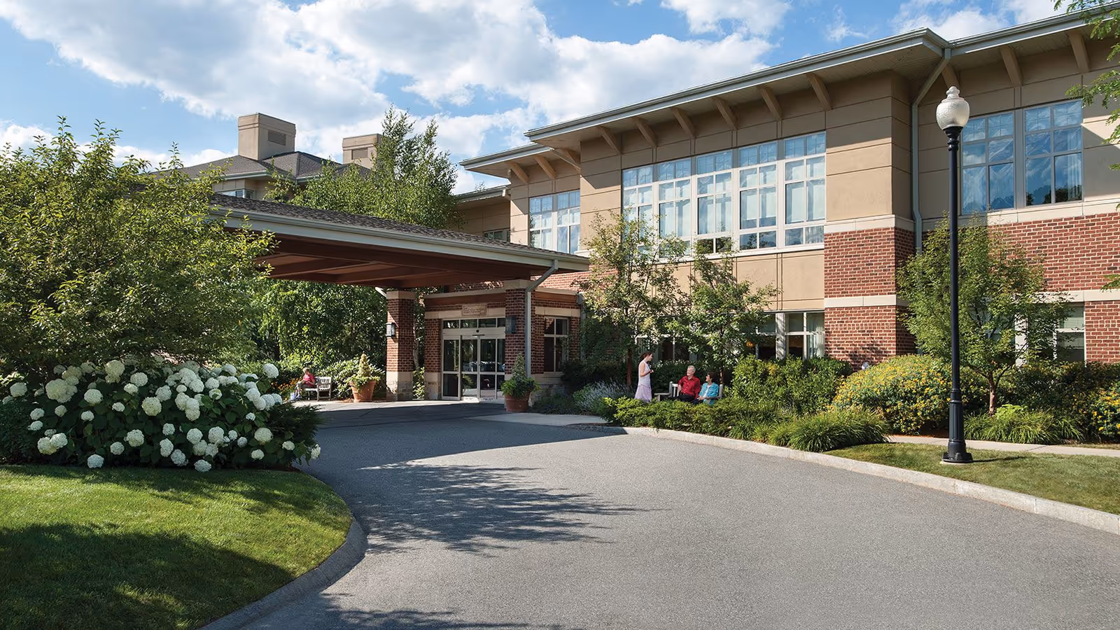 Exterior view of a senior living facility named Linden Ponds with a covered entrance, landscaped greenery, and a few people sitting and standing near the entrance on a sunny day.