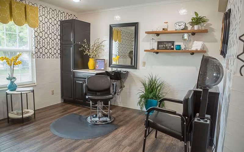 A well-lit hair salon room with a black salon chair in front of a mirror and a hair washing station. The room has wooden flooring, a window with patterned curtains, a small round table with a blue vase holding yellow flowers, and shelves with decorative items and towels. There is also a black chair with a hair dryer hood next to it and some green plants in blue pots.