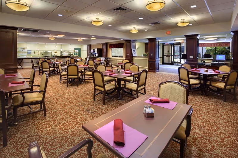 A spacious dining room with multiple wooden tables and cushioned chairs arranged neatly. Each table has a pink placemat and a folded red napkin. The room features patterned carpet flooring, warm lighting fixtures on the ceiling, and a serving area with a counter in the background. Large windows and glass doors allow natural light to enter the room.