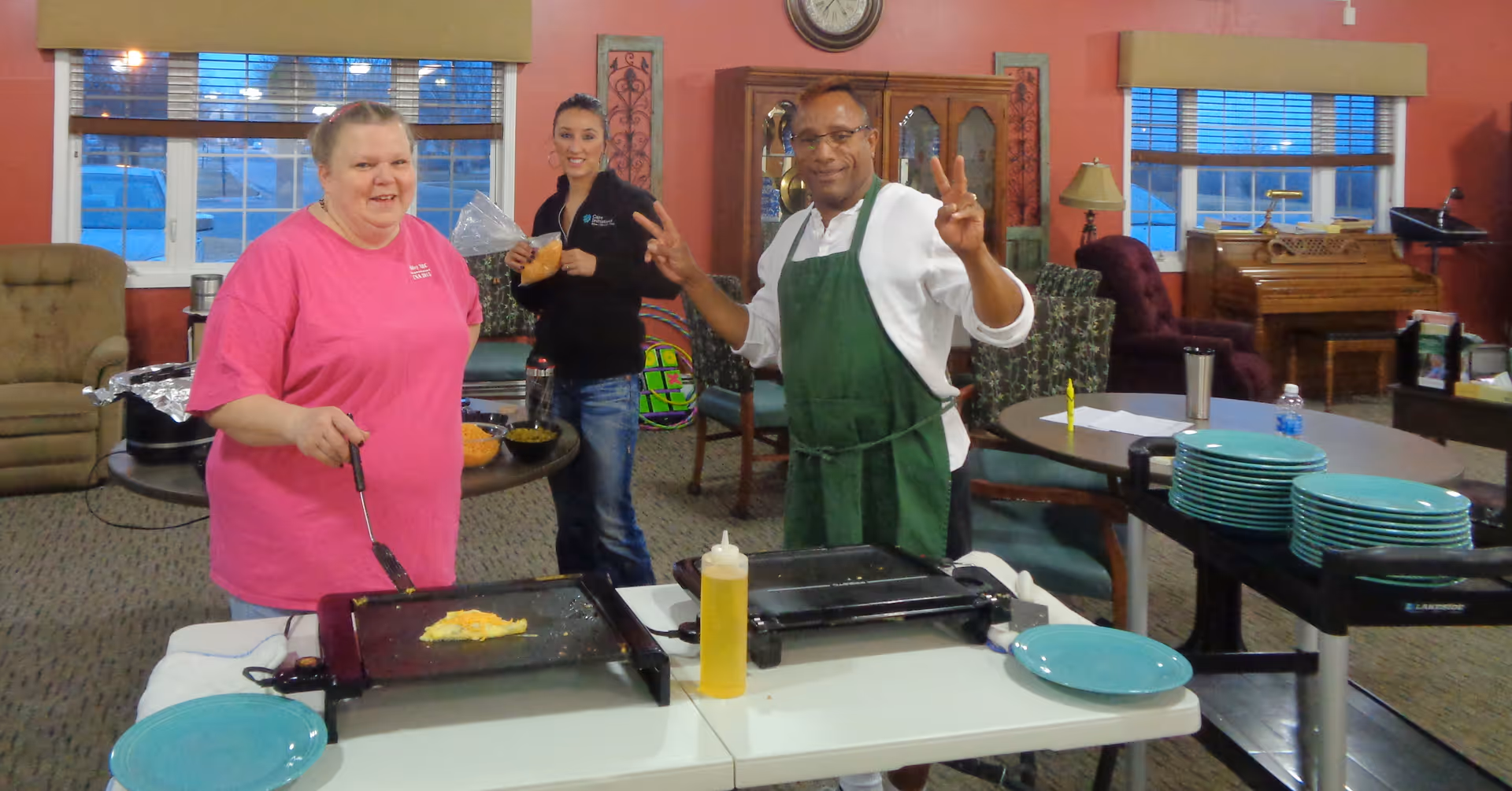 Three people in a common area of a senior living facility preparing food on electric griddles. One woman in a pink shirt is cooking, a man in a green apron is making a peace sign, and another woman in a black jacket is holding a drink. The room has tables, chairs, a piano, and large windows with blinds.