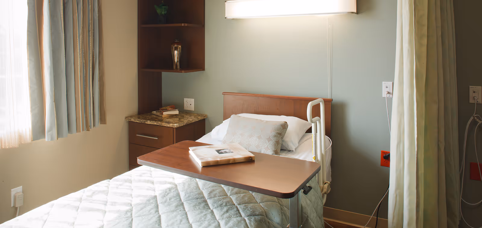 A single hospital-style bed with a light green quilted blanket and a decorative pillow, positioned next to a window with light curtains. A wooden overbed table holds a closed book. There is a small wooden cabinet with a granite top and shelves in the corner, and medical equipment outlets are visible on the wall.
