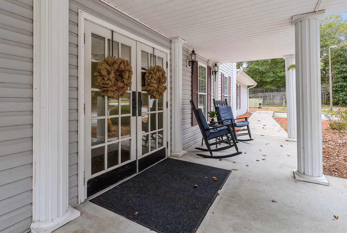 Covered porch area at the entrance of a building with two black rocking chairs with blue cushions, two white columns, double glass doors decorated with burlap wreaths, and a black doormat. There is a concrete walkway leading away from the porch and some greenery in the background.
