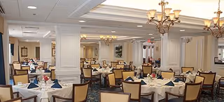 A spacious dining room with multiple round tables set with white tablecloths, blue folded napkins, and floral centerpieces. The room features beige walls, chandeliers, and cushioned chairs with wooden frames. Several people are seated at the tables in the background.
