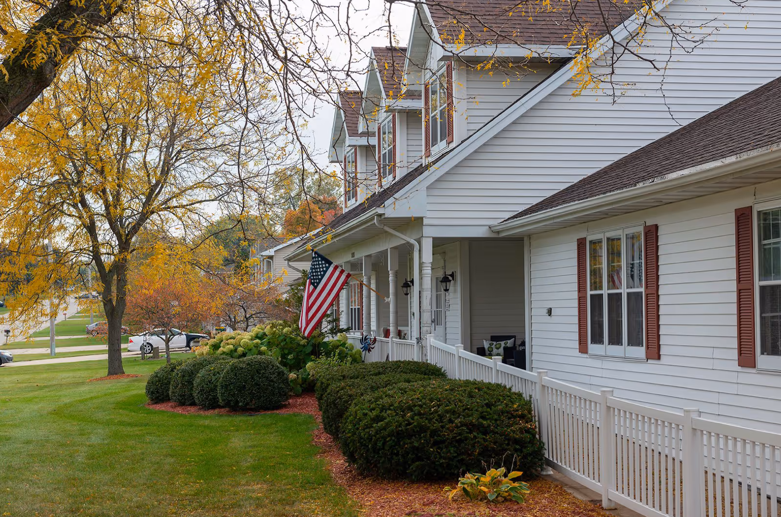Front view of a white-sided senior living building with a porch, American flag, manicured shrubs, and autumn trees.