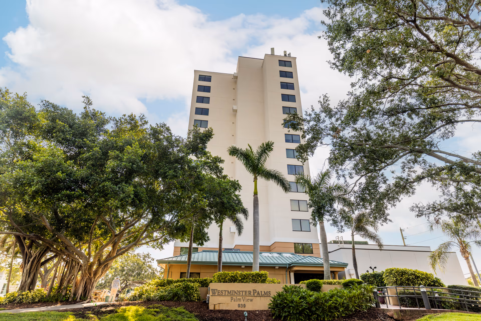 Tall multi-story building of Westminster Palms surrounded by trees and greenery under a partly cloudy sky.