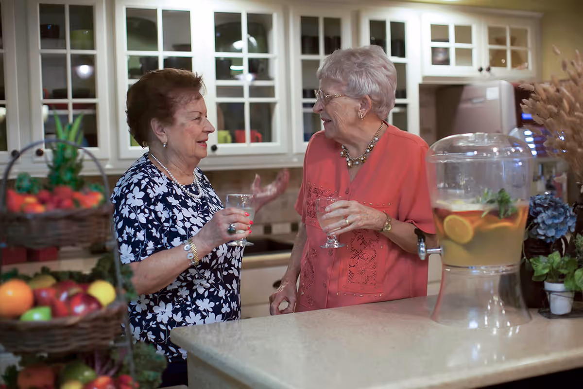 Two elderly women standing and talking in a kitchen area, each holding a glass of water. The kitchen has white cabinets with glass doors, a countertop with a large beverage dispenser containing lemon and herb infused water, and a basket of fresh fruits in the foreground.