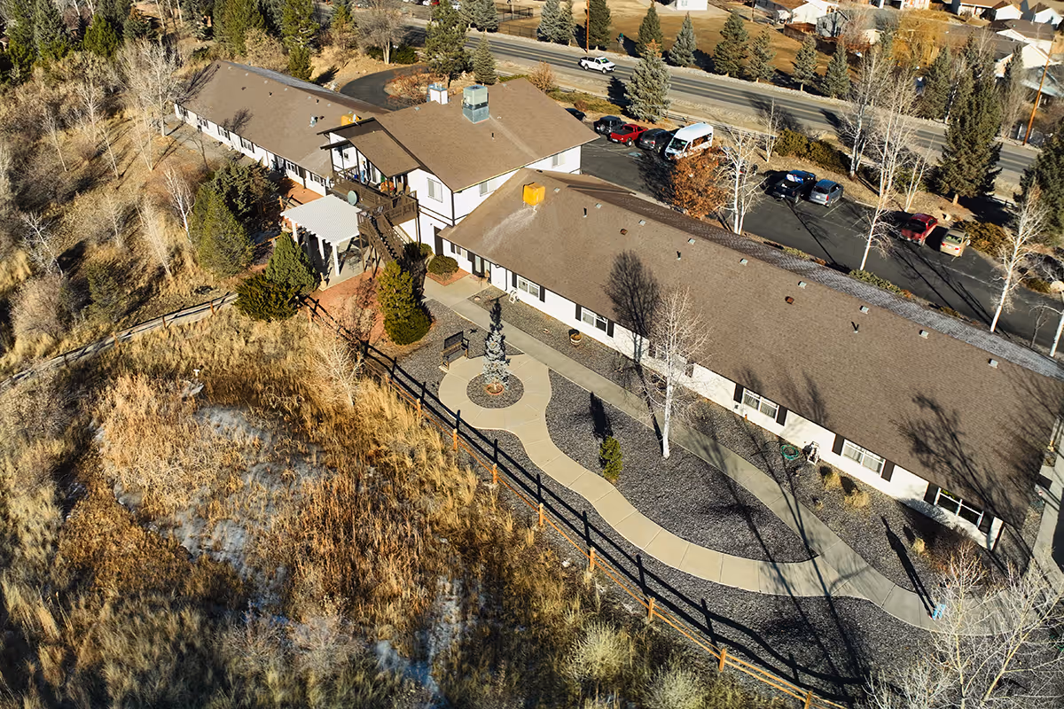 Aerial view of Heidi's Chateau, a senior living facility surrounded by trees and natural vegetation. The building has a brown roof and white exterior walls, with a paved walkway and a small circular garden area featuring a sculpture or fountain. Several cars are parked in the parking lot adjacent to the building.