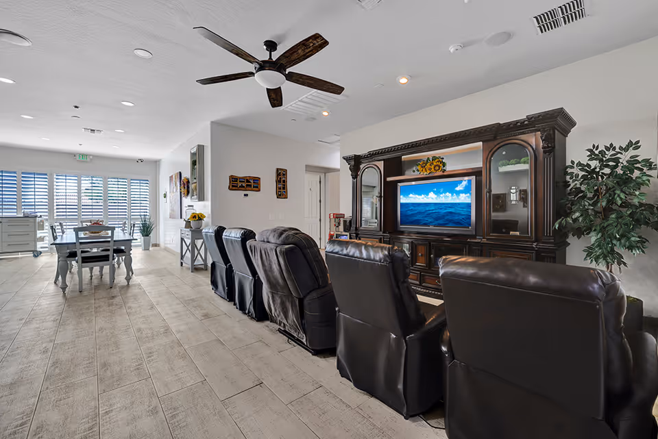 A spacious living area in an assisted living facility featuring a row of black leather recliners facing a large wooden entertainment center with a flat-screen TV displaying an ocean scene. To the left, there is a dining area with a table and chairs near large windows with white shutters, allowing natural light to fill the room. The floor is covered with light-colored wood-like tiles, and a ceiling fan is mounted above.