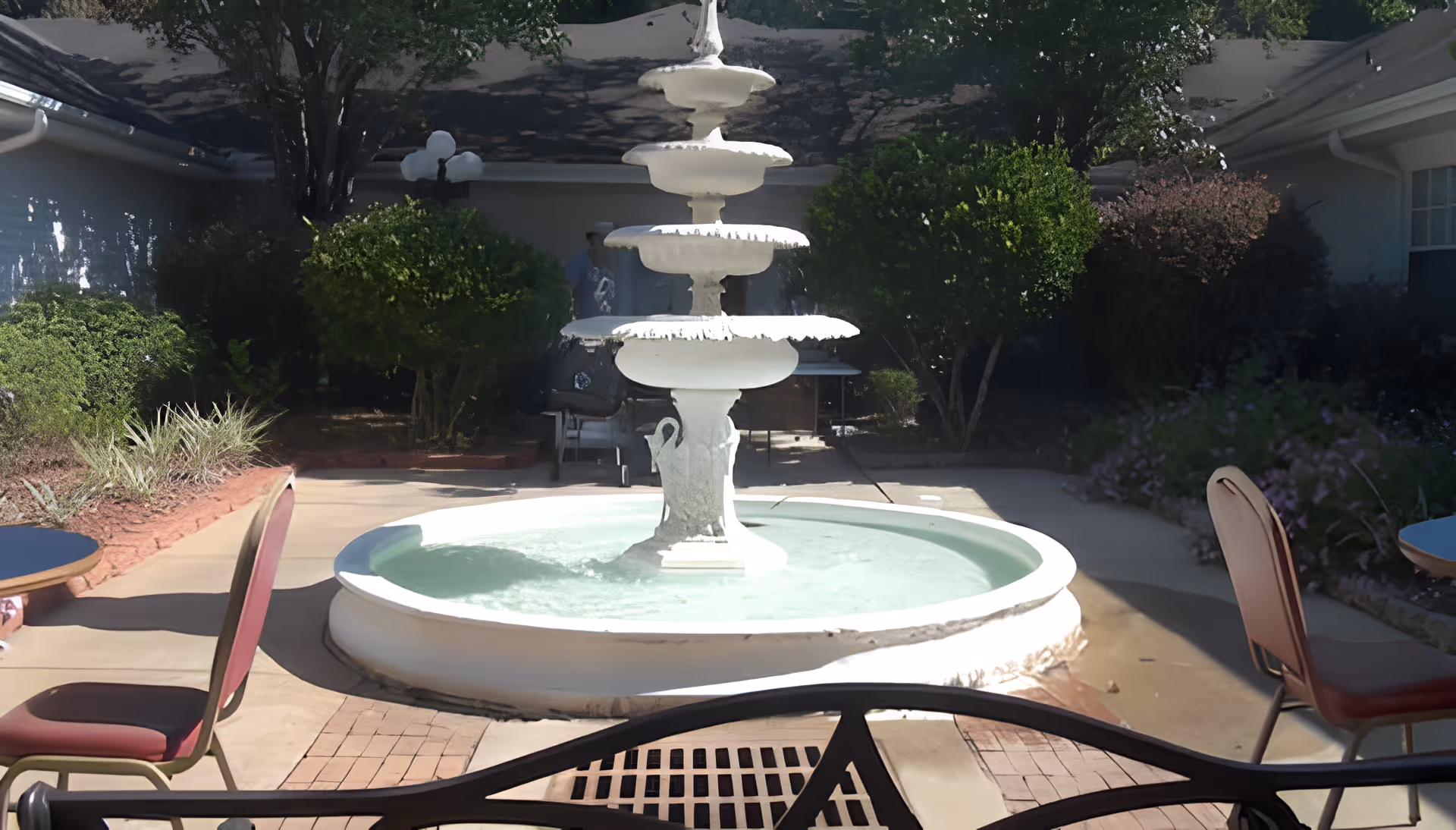 A multi-tiered white fountain in a sunlit courtyard with patio tables, chairs, and surrounding landscaping.