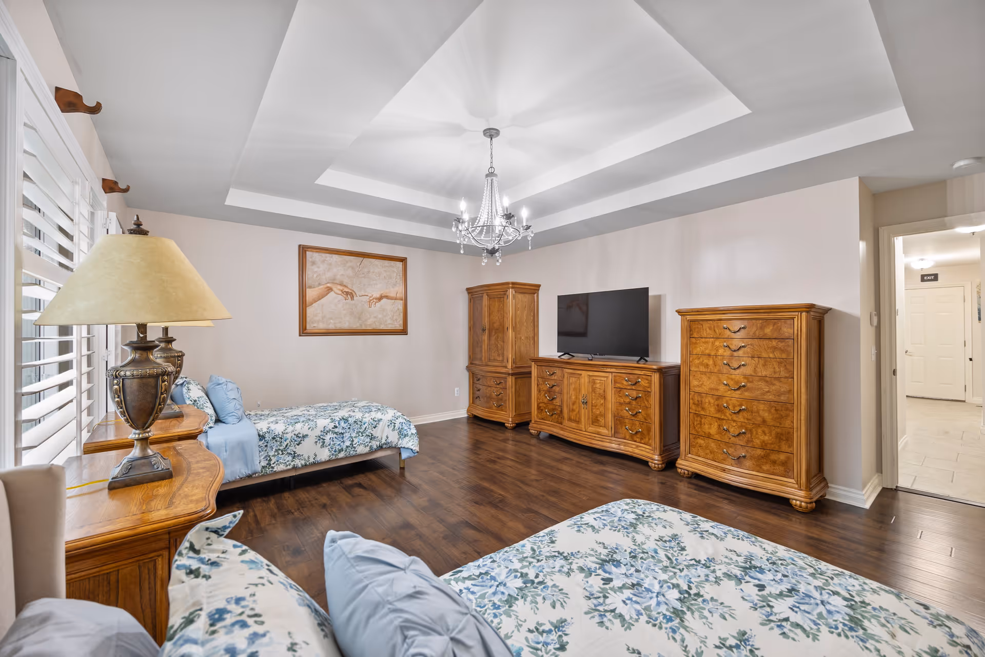 A spacious bedroom with two twin beds featuring floral bedspreads and blue pillows. The room has wooden furniture including a dresser, chest of drawers, and an armoire. A flat-screen TV sits on the dresser. A decorative chandelier hangs from a tray ceiling. There is a framed artwork on the wall depicting two hands reaching towards each other. A table lamp is on a wooden side table near the window with white plantation shutters. The floor is dark wood, and a doorway leads to a hallway with a visible exit sign.