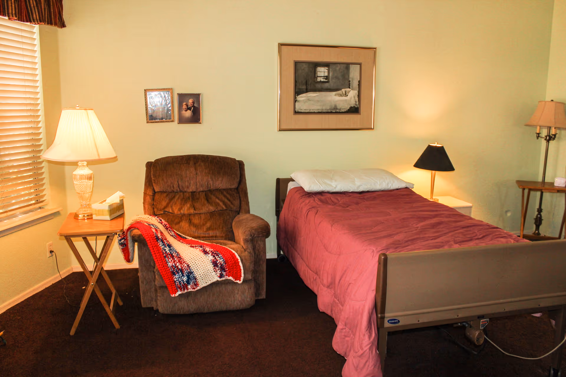 A cozy bedroom in an assisted living facility featuring a single bed with a pink comforter and a white pillow. Next to the bed is a brown recliner chair draped with a red, white, and blue knitted blanket. There are two side tables with lamps on either side of the bed. The walls are light-colored with framed pictures hanging above the chair and bed. A window with blinds is partially visible on the left side.
