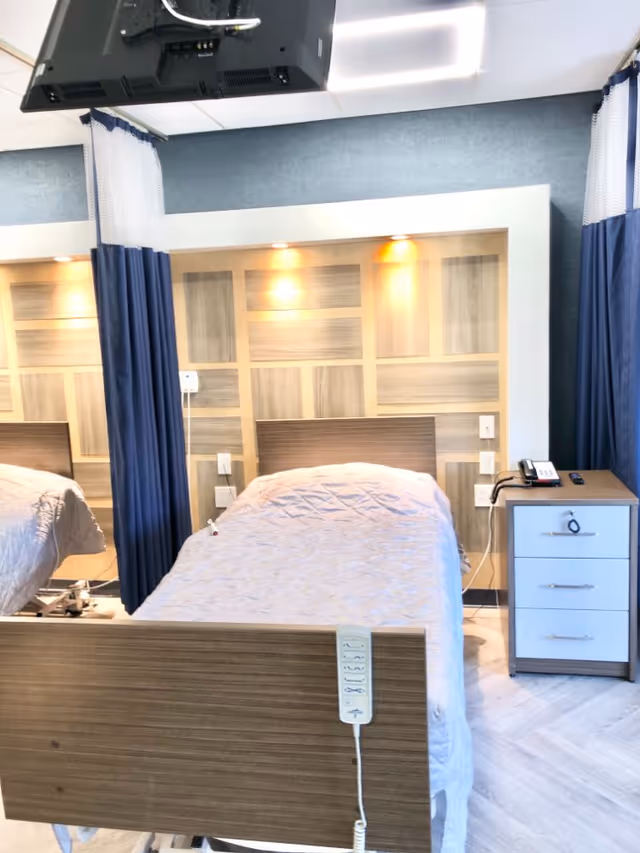 A hospital-style bed with a light gray quilted blanket in a room with wood paneling on the wall behind it. There are blue privacy curtains partially drawn on either side of the bed. A bedside table with a telephone and remote control is next to the bed. A ceiling-mounted television is visible above the bed.