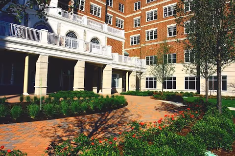 Outdoor courtyard area with brick-paved walkway, landscaped greenery, and trees in front of a multi-story brick and stone building with balconies and large windows.