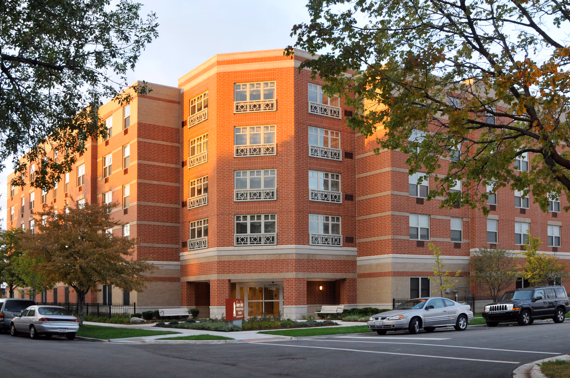 Exterior view of a multi-story brick senior living facility named Senior Suites of Marquette Village, with several windows, trees, parked cars, and a sidewalk in front.