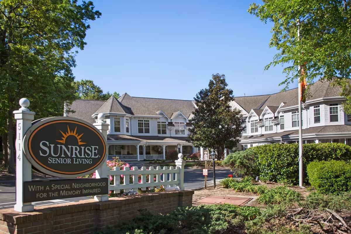 Exterior view of Sunrise Senior Living facility with a large sign in the foreground that reads 'Sunrise Senior Living with a special neighborhood for the memory impaired.' The building is two stories with white siding and multiple windows, surrounded by greenery and trees under a clear blue sky.