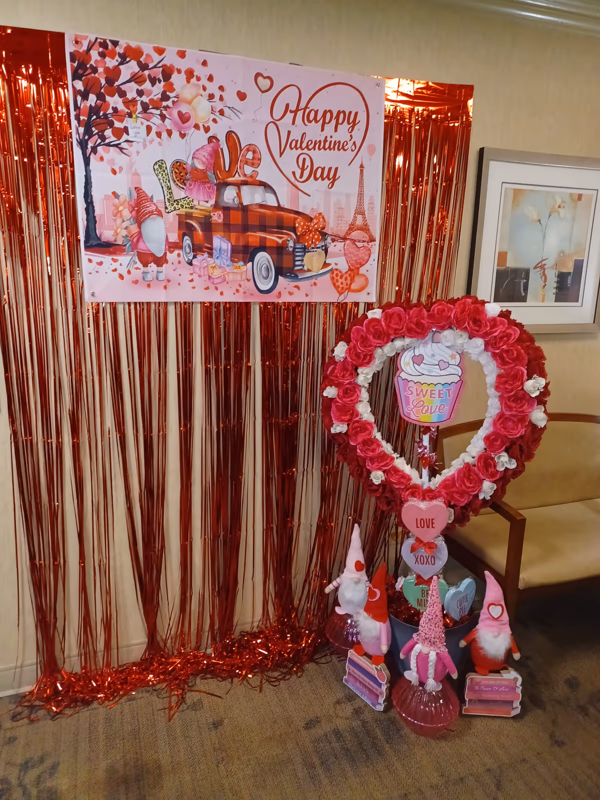 A Valentine's Day decoration setup inside a room with a red metallic fringe curtain backdrop. A poster with a red vintage truck, hearts, and the text 'Happy Valentine's Day' is displayed above. In front, there is a heart-shaped wreath made of red and white flowers with a cupcake decoration in the center that says 'Sweet Love'. Below the wreath, there are four small Valentine-themed gnome figurines holding signs with messages like 'Love', 'XOXO', and 'Be Mine'. A beige chair and a framed floral artwork are visible to the right.