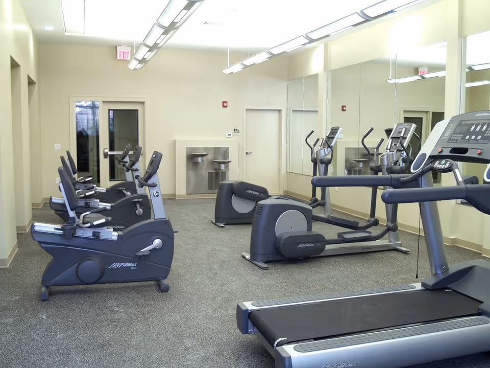 A fitness room with exercise equipment including stationary bikes, elliptical machines, and a treadmill. The room has beige walls, a carpeted floor, large mirrors on one wall, and fluorescent ceiling lights. There is a water fountain and two doors at the back of the room.