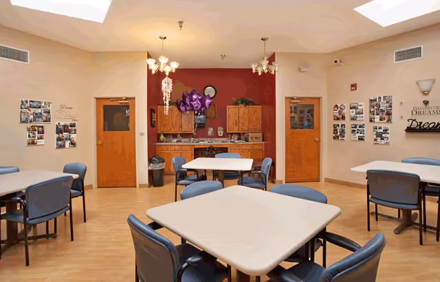 Communal dining/activity room with several square tables and chairs and a small kitchenette against a red accent wall.