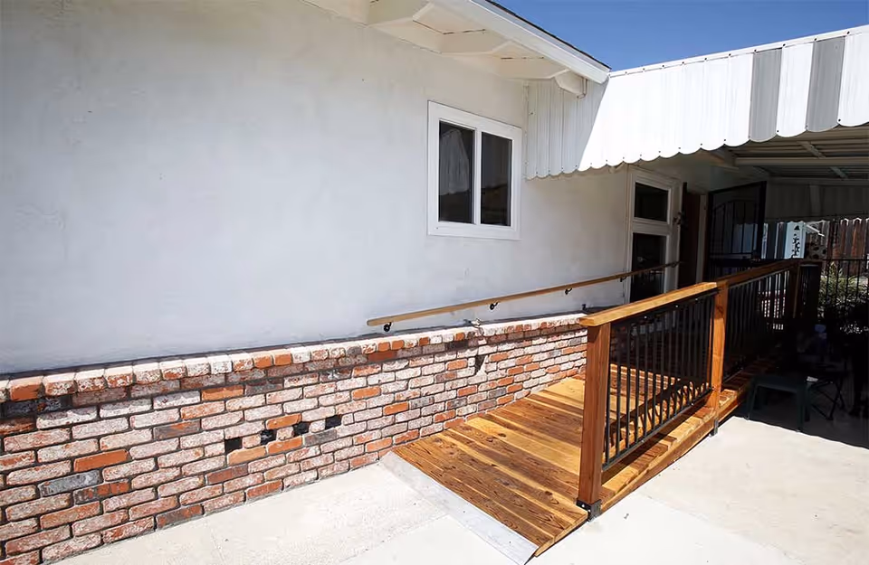 Wooden wheelchair ramp with metal balusters leading to a house entrance beside a brick half-wall and white stucco exterior under a metal awning.