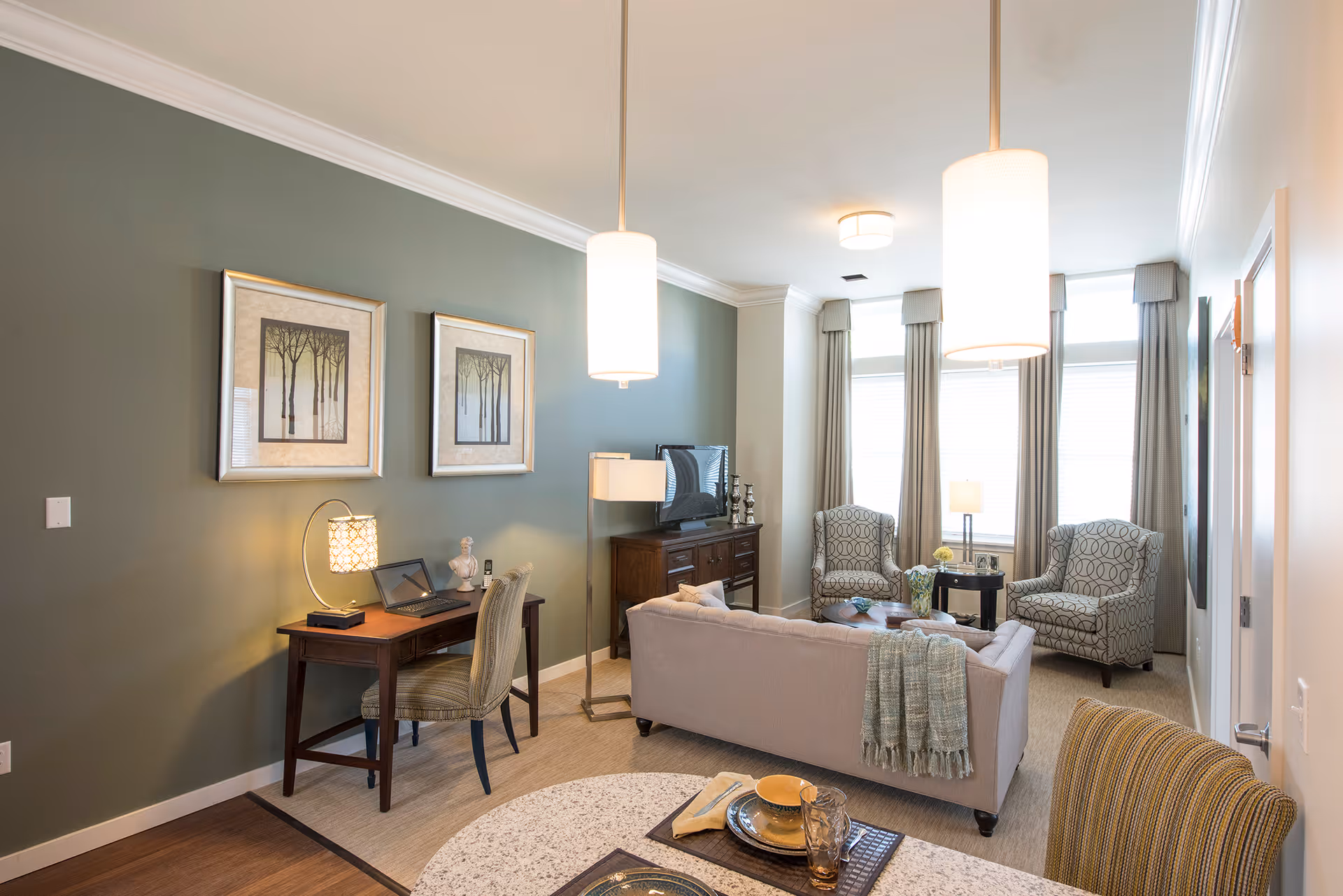 A cozy living room area with a beige sofa draped with a green throw blanket, two patterned armchairs near large windows with curtains, a wooden TV stand with a flat-screen TV, and a small wooden desk with a chair and a laptop. Two framed pictures of trees hang on a green accent wall. Two pendant lights hang from the ceiling, and a round dining table with place settings is partially visible in the foreground.