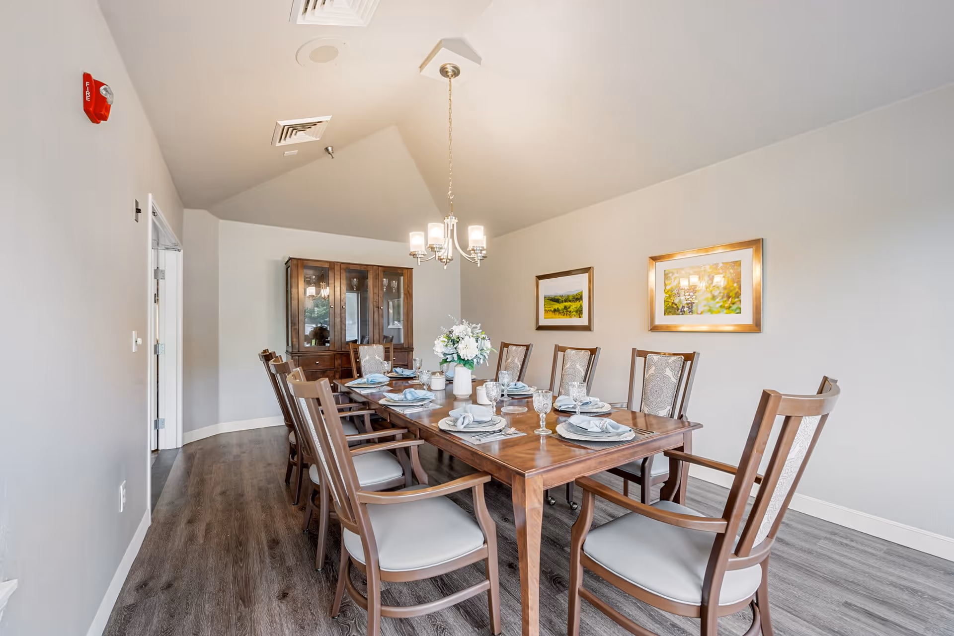 A dining room with a long wooden table set for eight people. The table is arranged with plates, napkins, glasses, and a floral centerpiece. There are eight wooden chairs with cushioned seats around the table. On the wall, there are two framed landscape paintings. A wooden cabinet with glass doors is visible in the background. The room has light-colored walls, wood flooring, and a chandelier hanging above the table.