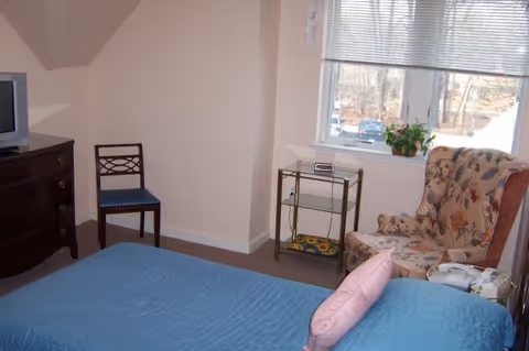 Bedroom with a blue-covered bed, floral armchair by a window, small side table, dresser with a TV and an extra chair.