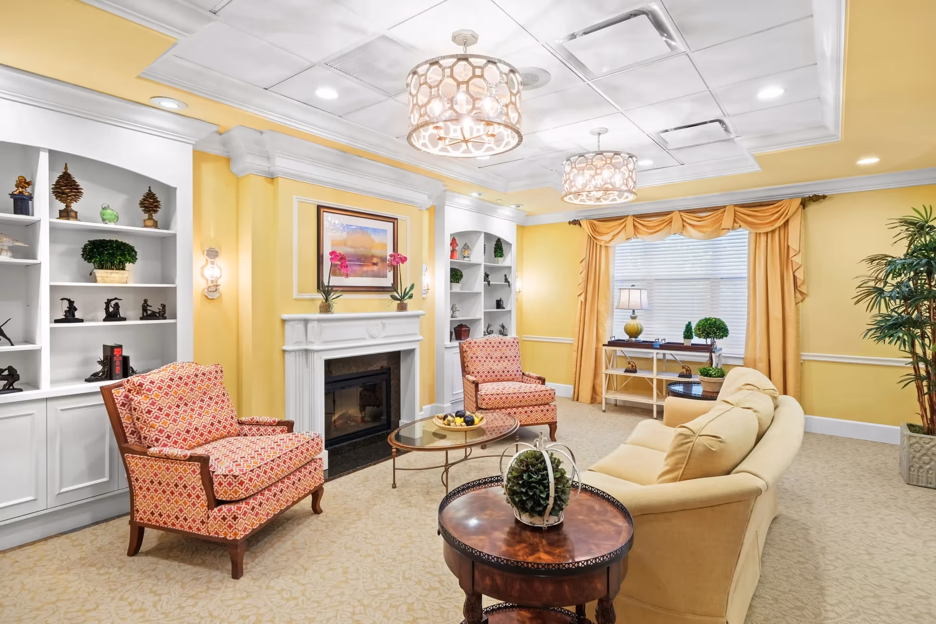 A cozy living room with yellow walls and carpeted floor featuring a beige sofa, two patterned armchairs, a white fireplace with a framed painting above it, built-in white shelves with decorative items, a round wooden coffee table with a small plant, and a window with yellow curtains. Two decorative ceiling lights hang from the white ceiling.