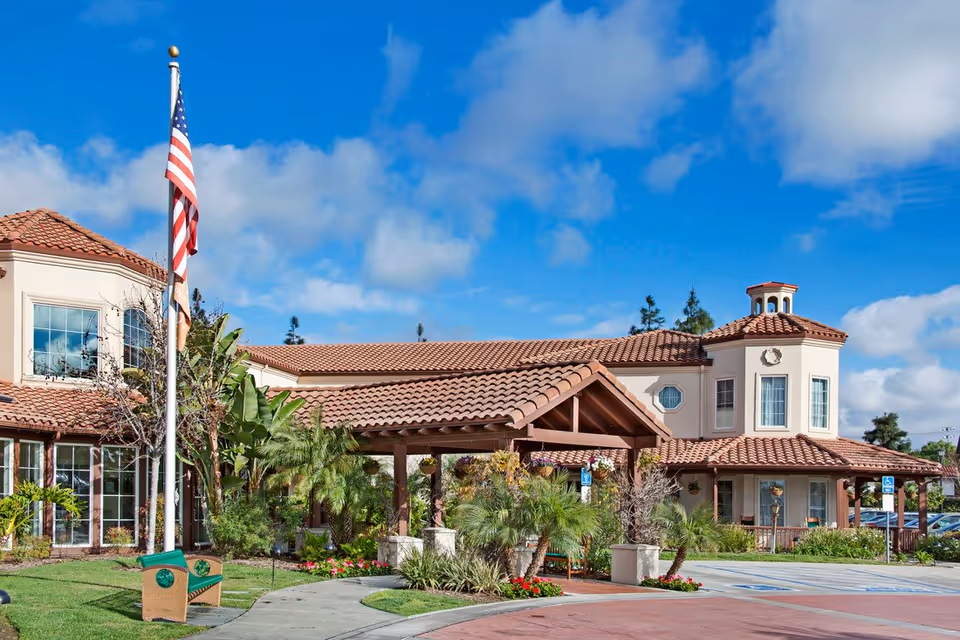 Exterior view of a senior living facility with a tiled roof, beige walls, and a covered entrance surrounded by lush greenery and palm trees. An American flag is flying on a flagpole near a green bench, with a clear blue sky and some clouds in the background.