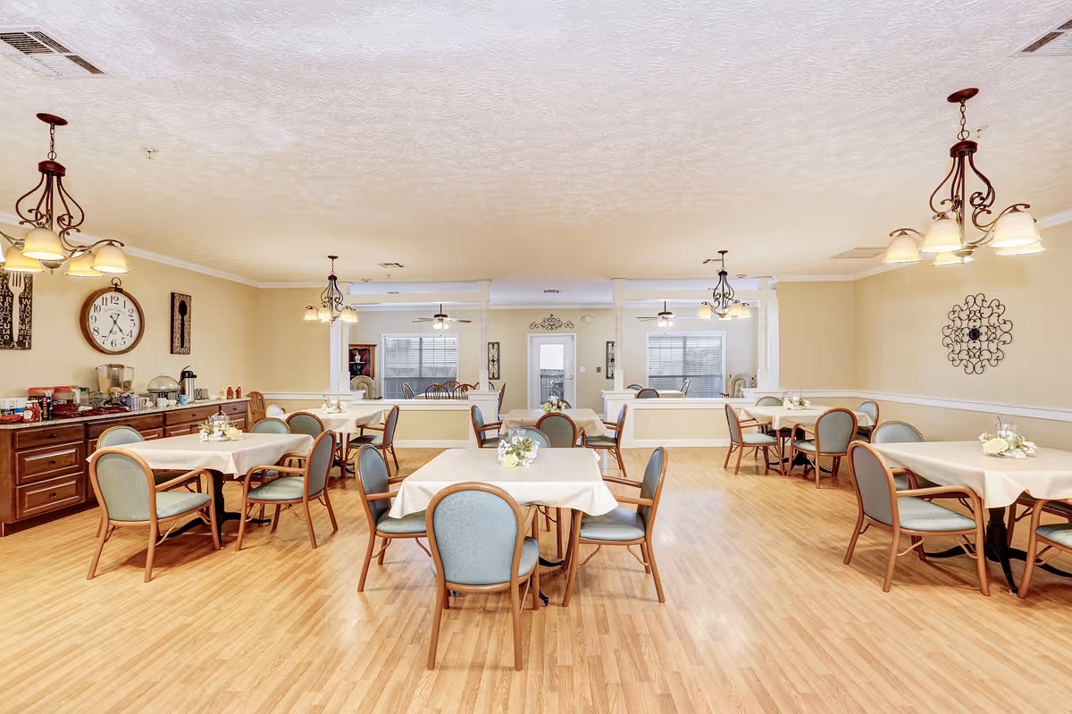 A bright and spacious dining room with several tables covered in white tablecloths, each surrounded by four chairs with light blue upholstery. The room features wooden flooring, beige walls, and multiple decorative light fixtures hanging from the ceiling. On the left side, there is a sideboard with various condiments and a large wall clock above it. The back wall has windows and a door, with additional seating visible in an adjoining room.